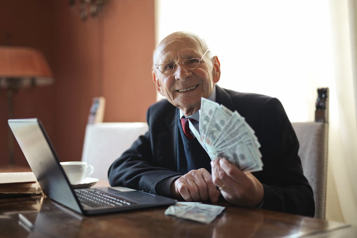 Papi souriant assis à son bureau tenant une liasse de billets en éventail derrières son ordinateur portable